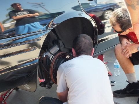 2003 Ford Crown Victoria - Mechanic crouches at the exposed front brake assembly of a black Ford as a seated man looks on.