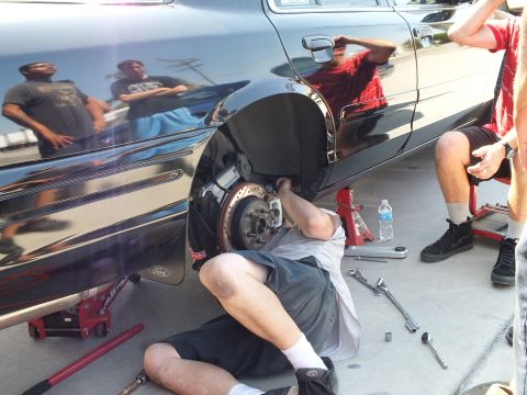 2003 Ford Crown Victoria - Person reclined at the front wheel well of a black Ford servicing the brakes, onlookers reflected in paint.