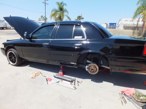 2003 Ford Crown Victoria - Black Panther sedan raised with hood up and front wheel off, hub and brakes exposed for service.