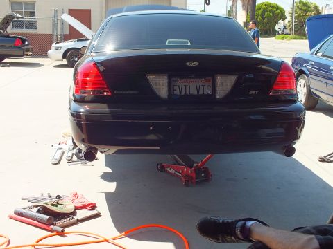 2003 Ford Crown Victoria - Black Ford rear with red taillights and dual exhaust lifted on a floor jack, plate reads EVIL VIC.