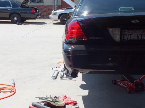 2003 Ford Crown Victoria - Tail of a black Ford raised for exhaust work with cut pipe sections staged on the ground.