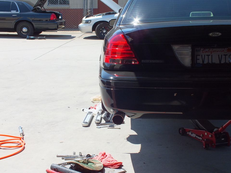 Rear of black Ford with vanity plate EVIL VIC and exhaust parts laid out on the shop floor