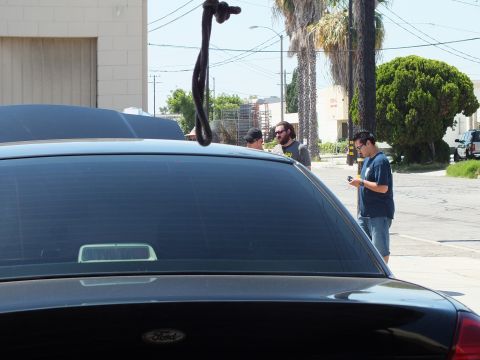 2003 Ford Crown Victoria - Trunk of a black Ford framing three men gathered in a palm-lined industrial street.