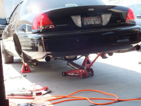 2003 Ford Crown Victoria - Rear of a black Ford lifted on stands and a 2.5-ton jack with a cut exhaust pipe hanging below.