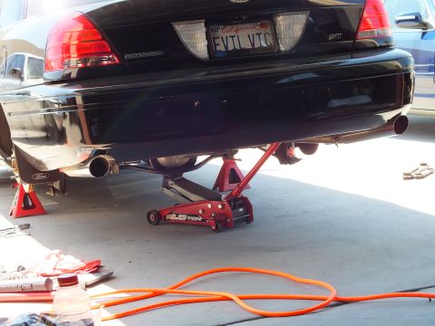 2003 Ford Crown Victoria - Black Panther sedan tail lifted on a floor jack with exhaust components exposed underneath.
