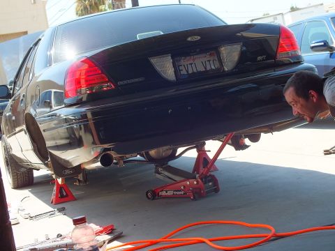 2003 Ford Crown Victoria - Person peers beneath the raised tail of a black Ford during exhaust work, orange air hose on floor.