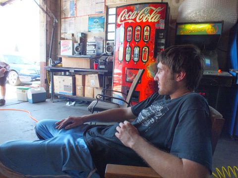 2003 Ford Crown Victoria - Man relaxes in a chair inside a cluttered garage with a Coca-Cola machine and arcade cabinet behind.