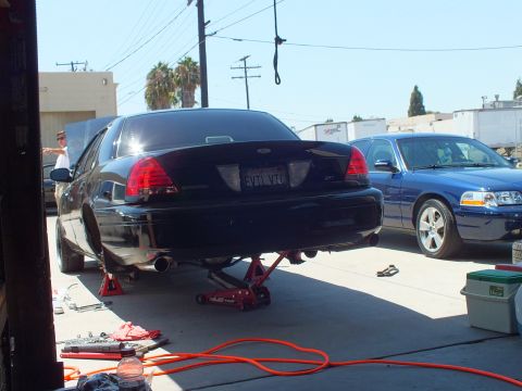 2003 Ford Crown Victoria - Rear three-quarter of a black Crown Victoria lifted on a floor jack, tools and air hose on the concrete, blue sedan nearby