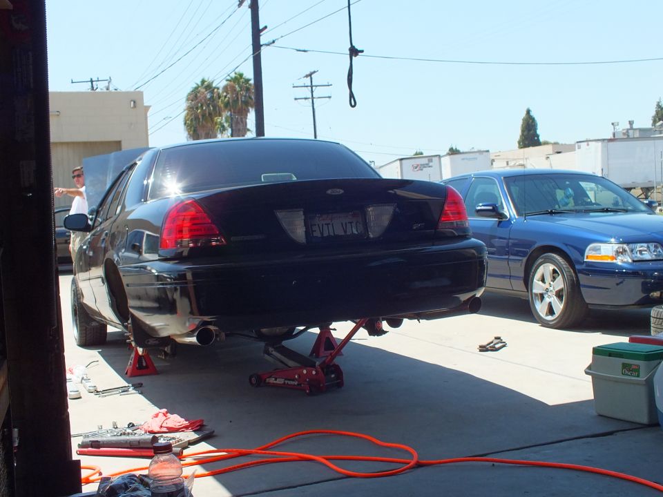 Black Ford Crown Victoria rear raised on red floor jack in shop driveway, blue sedan beside it