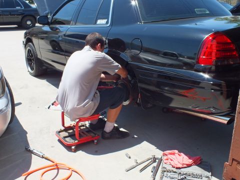 2003 Ford Crown Victoria - Mechanic kneeling on a creeper stool at the rear fender of a black sedan with red pinstripe flames, sockets laid out nearby