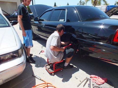 2003 Ford Crown Victoria - One man on a creeper works the rear fender of a black sedan with red flames while another stands watching by a silver van
