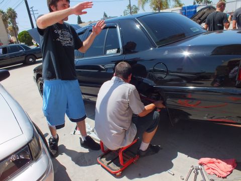 2003 Ford Crown Victoria - Standing man waves a hand while a seated mechanic works the rear quarter of a black sedan with red flame graphics