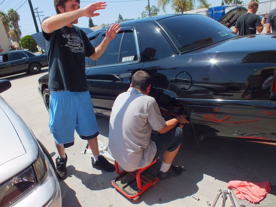 Man gesturing beside black flamed Ford Crown Victoria as another kneels on stool servicing the rear fender