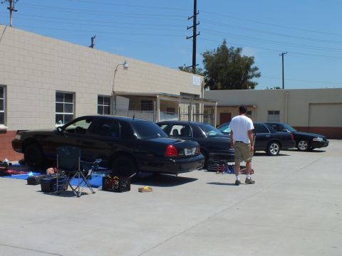 2003 Ford Crown Victoria - Several dark Panther-platform sedans lined up at a shop lot, folding chair and toolboxes set out, man walking past
