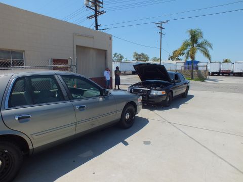 2003 Ford Crown Victoria - Gray sedan in foreground and a black sedan with its hood up, two people standing near the building