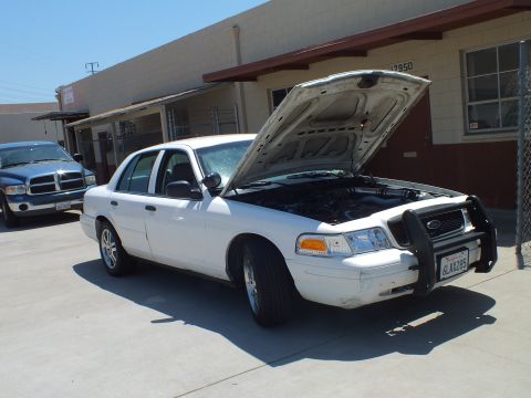 2003 Ford Crown Victoria - White Panther sedan with brush guard and raised hood beside a red Dodge Ram pickup outside a garage