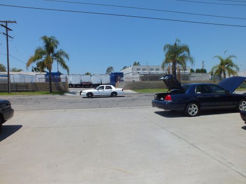 2003 Ford Crown Victoria - Distant white sedan on the road, foreground dark sedans with open trunk and hood, palms and rail cars behind