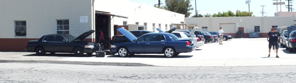 Panoramic row of dark Ford Crown Victoria sedans with open hoods outside a shop, man standing at right