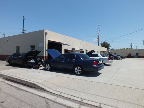 2003 Ford Crown Victoria - Two Panther sedans with raised hoods nose-out at a shop bay, more cars lined down the sunlit lot