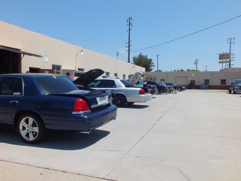 2003 Ford Crown Victoria - Rear of a blue sedan with deck spoiler leads a line of Panther cars, one hood up, parked at a shop lot