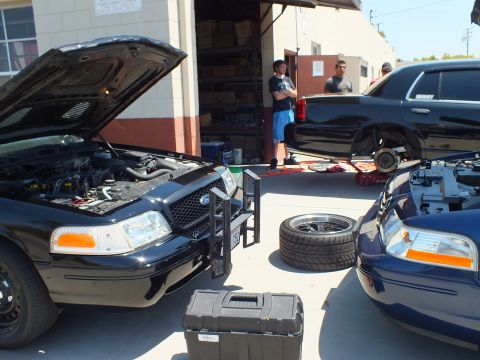 2003 Ford Crown Victoria - Two black Panther sedans with open hoods at a shop bay, a wheel and large toolbox between them, men inside