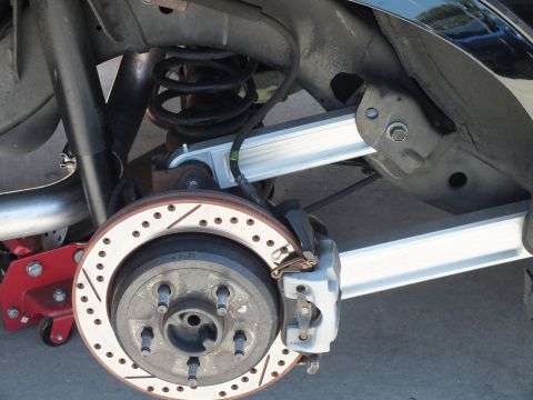 2003 Ford Crown Victoria - Underside view of a rear disc brake setup, drilled rotor, coil spring and aluminum lateral links over a red jack