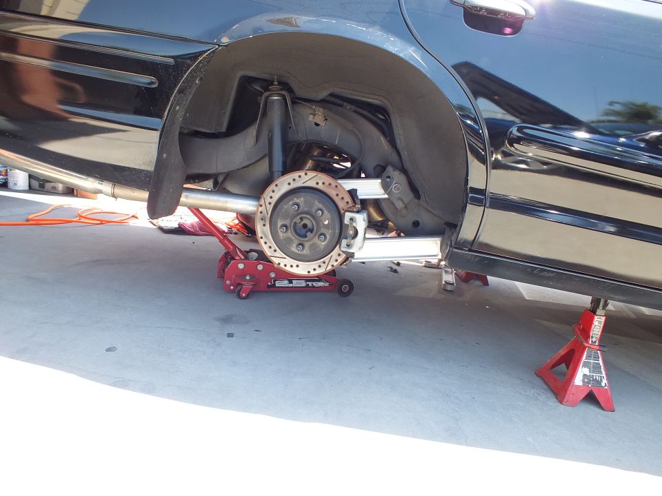 Black sedan raised on red floor jack with rear brake rotor and aluminum control arms exposed