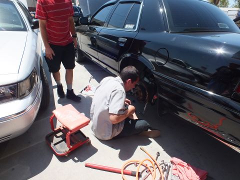 2003 Ford Crown Victoria - Technician crouched beside a black sedan with a creeper seat and tools, working the rear brakes outdoors