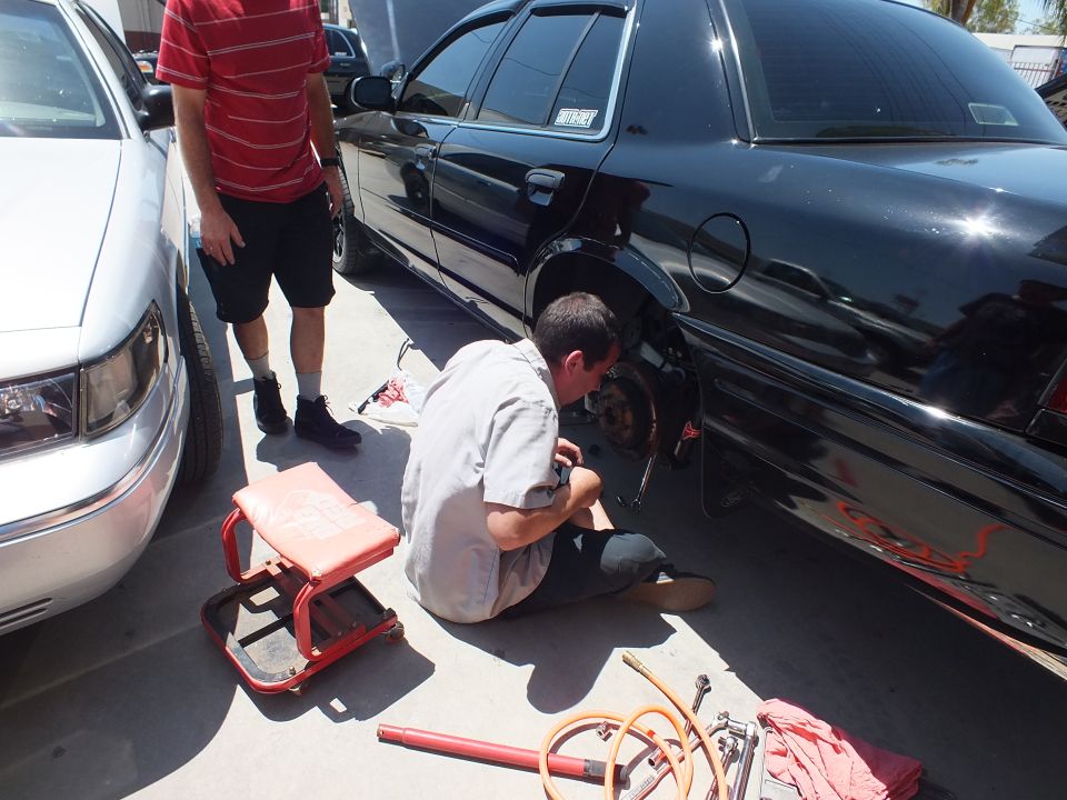 Man kneeling at rear wheel of black sedan working on brakes in industrial parking lot