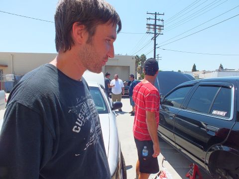 2003 Ford Crown Victoria - Man in dark shirt in foreground, second man in red holds a water bottle near parked sedans on jack stands