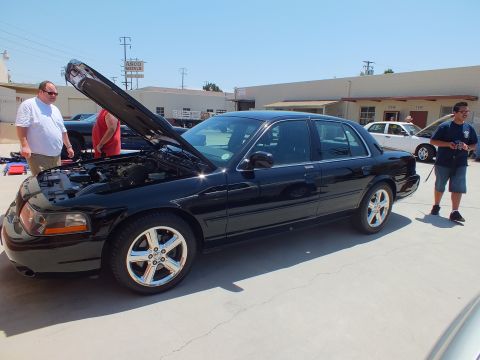 2003 Ford Crown Victoria - Black full-size sedan, hood raised, polished five-spoke wheels, parked by an ASCO Metals building