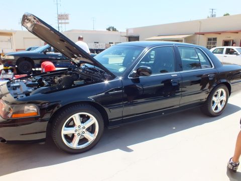 2003 Ford Crown Victoria - Front three-quarter of a black sedan, hood open and chrome wheels, with a gold sedan in the background