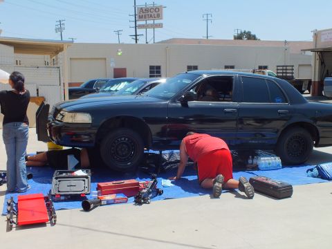 2003 Ford Crown Victoria - Black sedan lifted on jacks with bare steel rotors, tarps and red tool chests spread across the lot