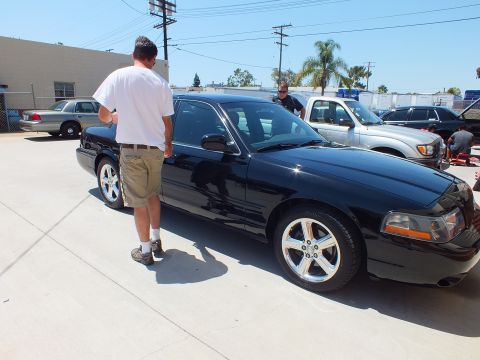 2003 Ford Crown Victoria - Man views a black sedan with polished five-spoke wheels, other Panther sedans parked behind