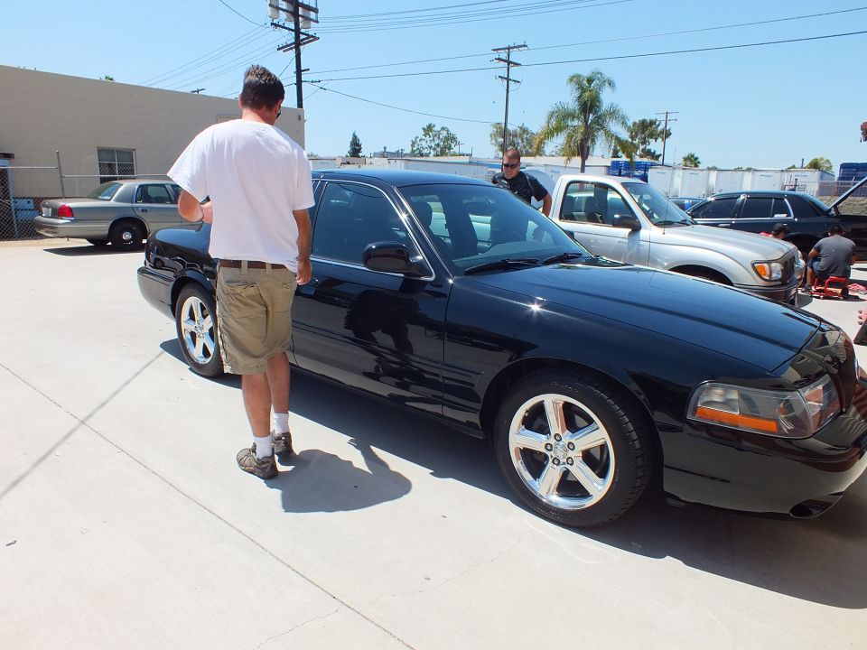 Man in white shirt standing beside black sedan with chrome five-spoke wheels in parking lot