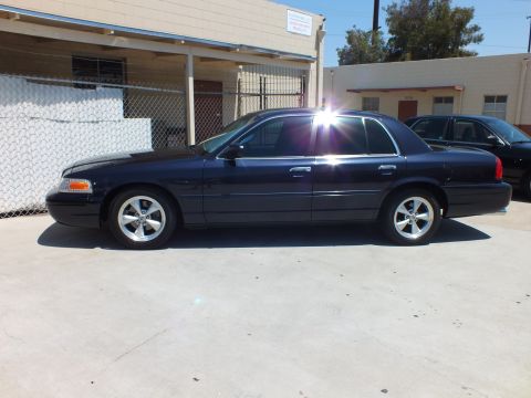 2003 Ford Crown Victoria - Side profile of a dark blue Crown Victoria with chrome wheels, sun glare on the roof, industrial backdrop