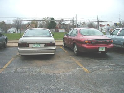 1997 Ford Taurus - Rear view of a red four-door sedan with rounded oval taillights parked beside a beige sedan in a paved lot, both facing away from the camer…