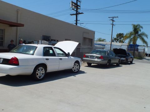 2003 Ford Crown Victoria - White Crown Victoria, hood raised, alongside a gray and a black sedan against a warehouse wall