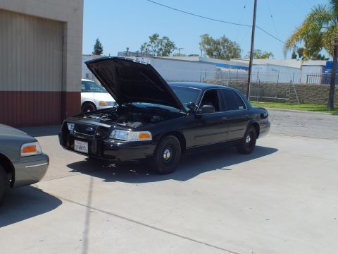 2003 Ford Crown Victoria - Black Crown Victoria with hood up and steel wheels parked near a roll-up door, front three-quarter view