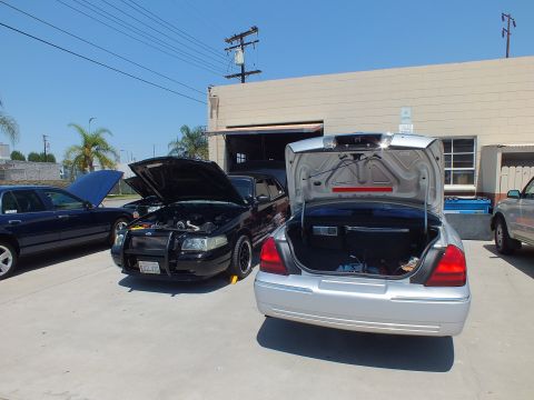 2003 Ford Crown Victoria - Black Ford sedan with brush guard beside a silver sedan with raised trunk lid at an open warehouse door
