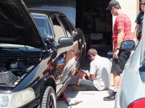 2003 Ford Crown Victoria - Group of men around a lifted black sedan, one seated writing on a red case, side mirror in foreground