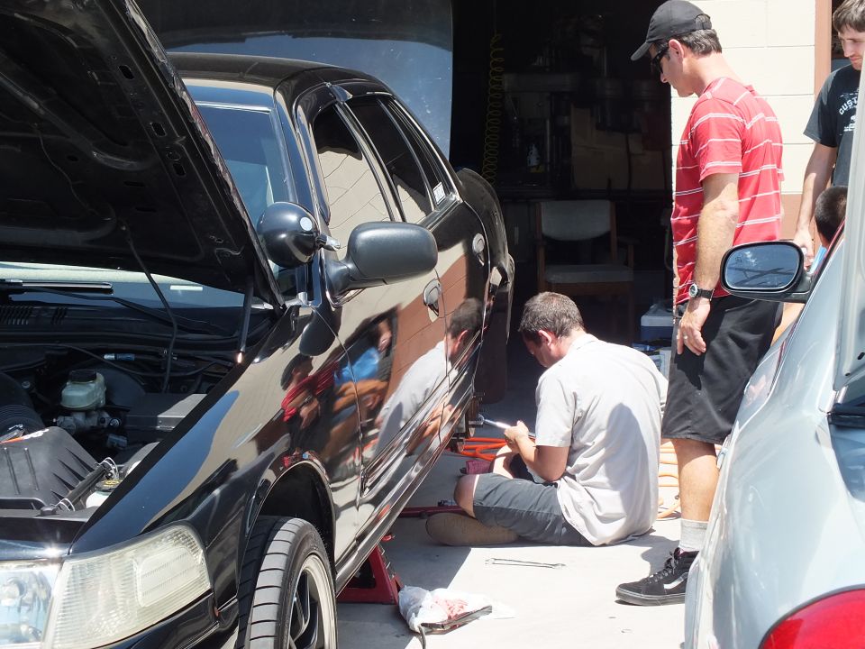 Men gathered around black sedan on jack as one kneels writing on red toolbox in lot