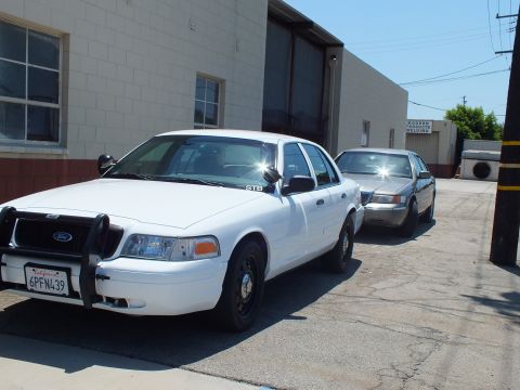 2003 Ford Crown Victoria - White Crown Victoria with a brush guard and plate 6PFN439 parked ahead of a gray sedan by a warehouse