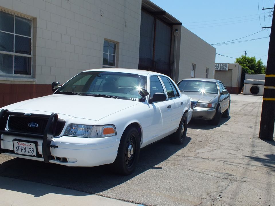 White Ford Crown Victoria with push bar and gray sedan parked along warehouse driveway