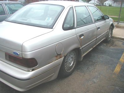 1997 Ford Taurus - Rear three-quarter view of a silver second-generation Ford Taurus sedan in a parking lot, showing rust bubbling around the rear wheel arch…