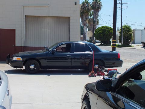 2003 Ford Crown Victoria - Black Ford Crown Victoria sits with its front fascia off, jack and tools on the ground beside a roll-up shop door.