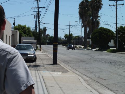 2003 Ford Crown Victoria - man stands curbside as a dark coupe drives off down a palm-lined street, a silver Mercury parked nearby.