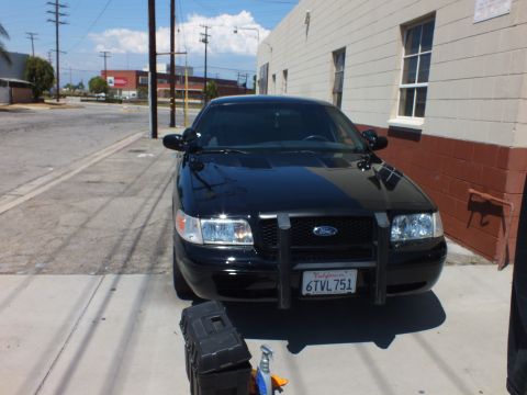2003 Ford Crown Victoria - Black Ford Crown Victoria front end with a push bumper and California plate 6TVL751, parked beside a building.