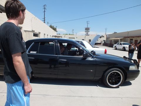 2003 Ford Crown Victoria - man looks on as a lowered black Mercury Grand Marquis on black wheels sits in a shop lot, hood ajar.
