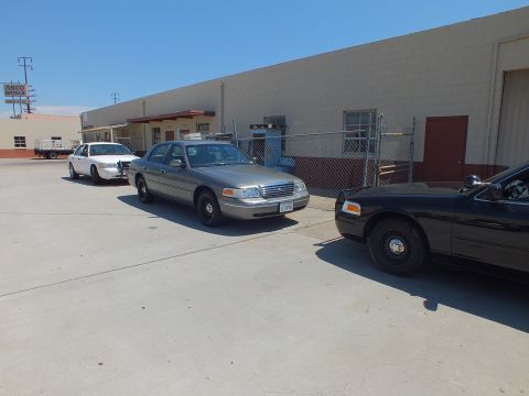 2003 Ford Crown Victoria - Three Ford Crown Victoria sedans in white, tan and black parked by a fenced industrial yard.